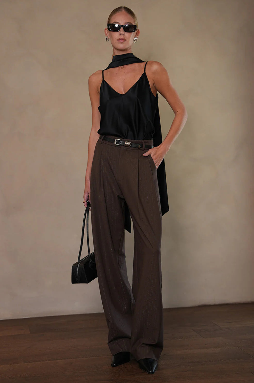 Woman wearing a black sleeveless top and brown wide-leg pants, holding a black handbag against a beige background. tailored trousers.