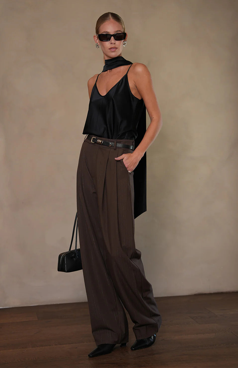 Woman in black halter top and brown wide-leg pants holding a black handbag against a neutral background